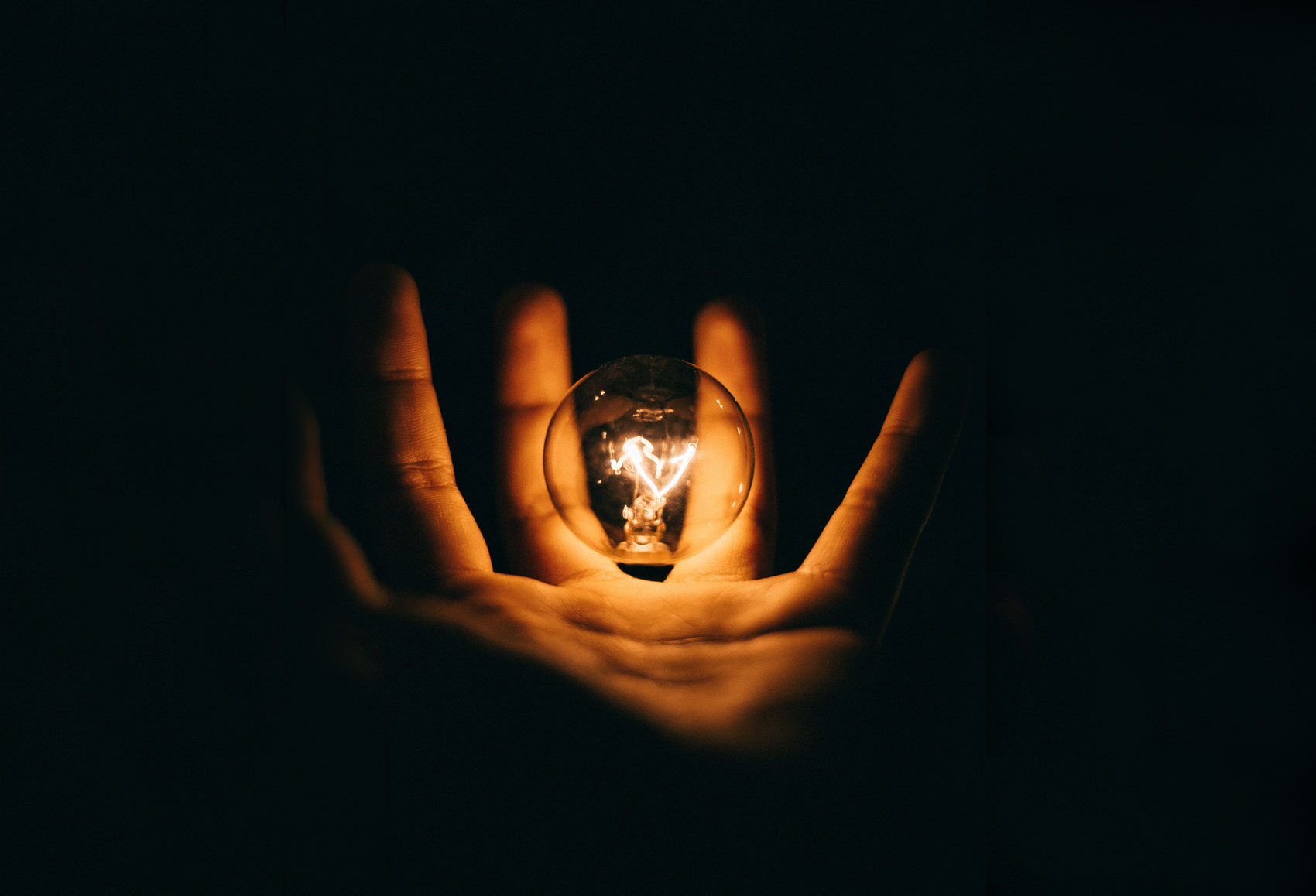 Eine Hand hält eine leuchtende Glühbrine. Der Lichtschein reflektiert auf der Handinnenseite, der Rest liegt im Dunkeln
