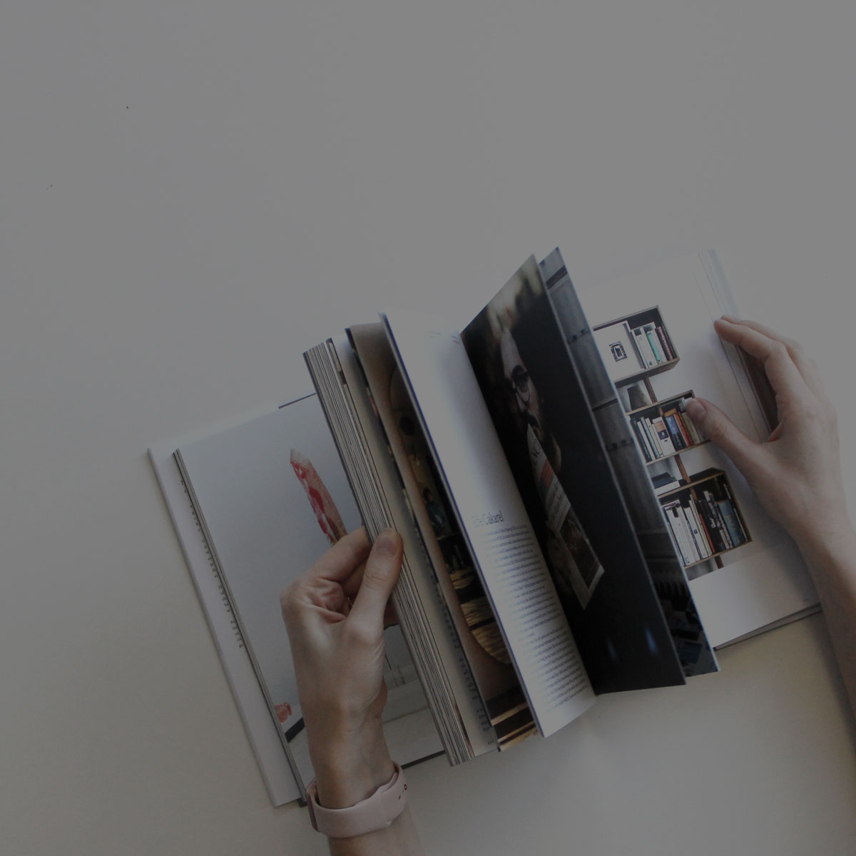 Eine Frau blättert in einem Buch. Die Darstellung aus der Vogelperspektive ist sehr reduziert auf die Hände und das Buch. Das Buch liegt auf einem weißen Tisch. Das ganze Bild ist leicht verdunkelt.